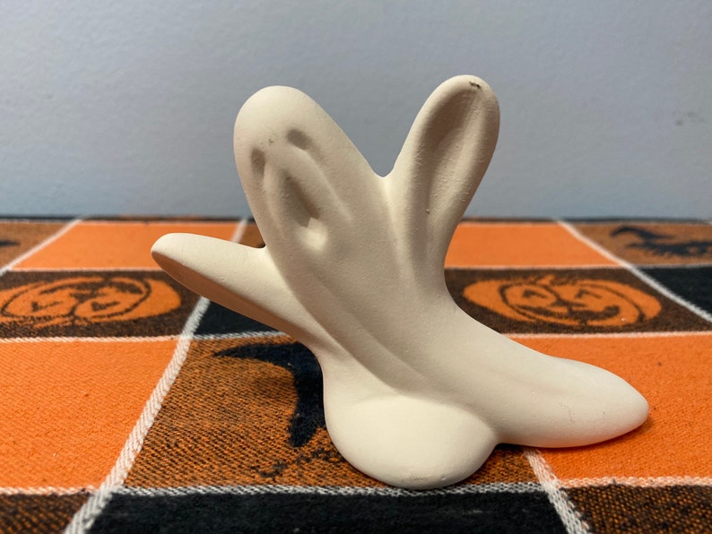 Puede incluir: Una figura de fantasma de cer&aacute;mica de color crema claro. El fantasma tiene una cara simple y amigable y los brazos extendidos. La figura est&aacute; sobre un mantel a cuadros naranja y negro con tem&aacute;tica de Halloween.