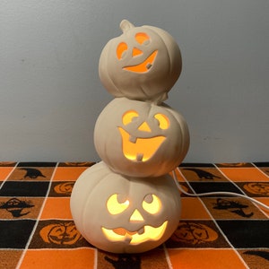 May include: A stacked ceramic Halloween decoration featuring three carved pumpkin faces with illuminated interiors. The pumpkins are white and sit on a black and orange checkered tablecloth. A power cord extends from the base.