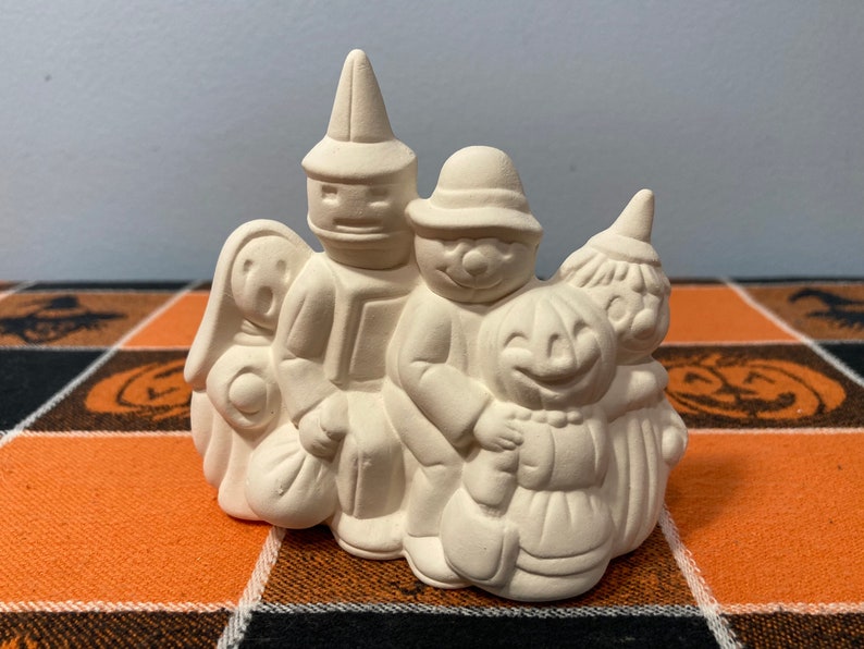 Puede incluir: Una figura de cer&aacute;mica de Halloween que representa un grupo de figuras disfrazadas. Las figuras incluyen un fantasma, un mago, un ni&ntilde;o con sombrero, una calabaza y una bruja. La figura no est&aacute; pintada y est&aacute; lista para personalizar.