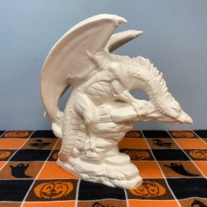 Peut inclure: Figurines de dragon en céramique blanche avec des ailes déployées, perchées sur une formation rocheuse. Le dragon est en position accroupie, la tête tournée sur le côté.