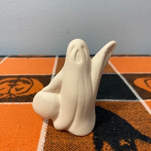 Puede incluir: Peque&ntilde;a figura de fantasma de cer&aacute;mica sin pintar, con los brazos extendidos y un objeto redondo en la mano izquierda. El fantasma es blanco y est&aacute; sobre un mantel a cuadros naranja y negro con tem&aacute;tica de Halloween.