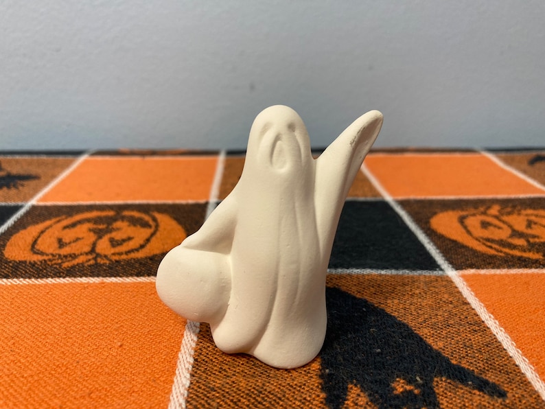 Puede incluir: Peque&ntilde;a figura de fantasma de cer&aacute;mica sin pintar, con los brazos extendidos y un objeto redondo en la mano izquierda. El fantasma es blanco y est&aacute; sobre un mantel a cuadros naranja y negro con tem&aacute;tica de Halloween.