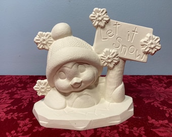 Może przedstawiać: Biała ceramiczna figurka pingwina w czapce, trzymająca tabliczkę z napisem "Let it Snow". Pingwin siedzi na podstawie przypominającej lód.