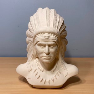 May include: White ceramic bust of a Native American man wearing a headdress. The bust is detailed with feathers and a beaded necklace.