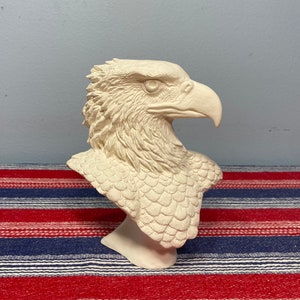 May include: A white ceramic sculpture of an eagle's head and neck. The eagle has a detailed, textured surface and a sharp beak.