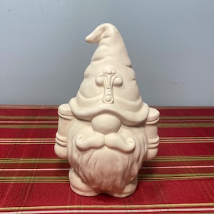 May include: A ceramic gnome figurine in an off-white color. The gnome has a large hat with a decorative emblem, a long white beard, and holds two cylindrical objects. The figurine is on a red and green striped surface.