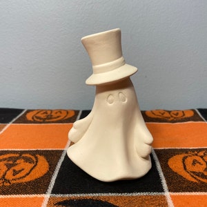 May include: White ceramic ghost figurine wearing a top hat. The ghost has two small circles for eyes. The figurine is sitting on a black and orange checkered fabric.