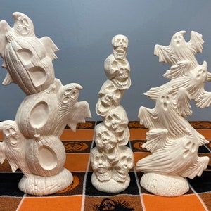 May include: Three ceramic Halloween decorations, each featuring a different spooky theme. One is a stack of pumpkins with ghosts, another is a stack of skulls, and the last is a stack of ghosts.