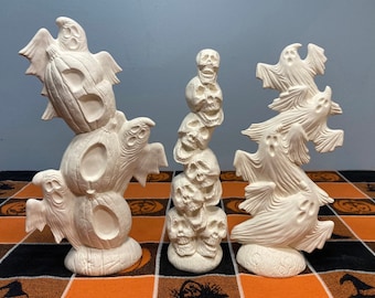 May include: Three ceramic Halloween decorations, each featuring a different spooky theme. One is a stack of pumpkins with ghosts, another is a stack of skulls, and the last is a stack of ghosts.