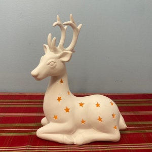 May include: A white ceramic deer figurine with star-shaped cutouts. The deer is in a resting position, with its antlers pointing upwards. The star cutouts allow light to shine through, creating a warm glow. The figurine is on a red and green striped surface.