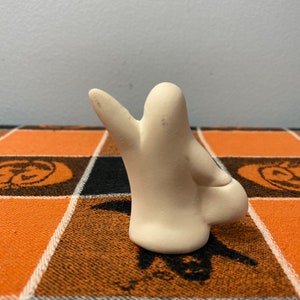 Puede incluir: Peque&ntilde;a figura de fantasma de cer&aacute;mica blanquecina con una mano levantada. El fantasma est&aacute; colocado sobre un mantel a cuadros naranja y negro con tem&aacute;tica de Halloween, con dise&ntilde;os de calabazas y murci&eacute;lagos.