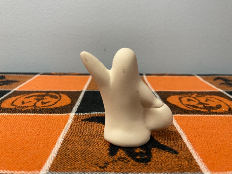 Puede incluir: Peque&ntilde;a figura de fantasma de cer&aacute;mica blanquecina con una mano levantada. El fantasma est&aacute; colocado sobre un mantel a cuadros naranja y negro con tem&aacute;tica de Halloween, con dise&ntilde;os de calabazas y murci&eacute;lagos.