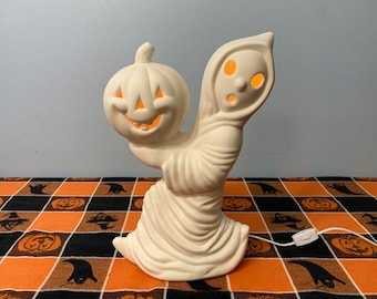 May include: White ceramic ghost holding a white ceramic pumpkin with a jack-o'-lantern face. Both the ghost and pumpkin have orange lights inside.
