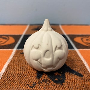 Puede incluir: Una peque&ntilde;a calabaza de cer&aacute;mica sin pintar con una cara sonriente. La calabaza tiene un tallo y est&aacute; sobre una tela a cuadros naranja y negra con dise&ntilde;os tem&aacute;ticos de Halloween.
