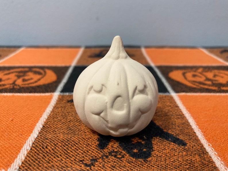 Puede incluir: Una peque&ntilde;a calabaza de cer&aacute;mica sin pintar con una cara sonriente. La calabaza tiene un tallo y est&aacute; sobre una tela a cuadros naranja y negra con dise&ntilde;os tem&aacute;ticos de Halloween.