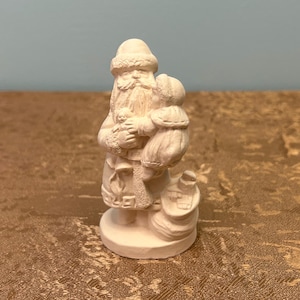 May include: A small, unpainted ceramic Santa Claus figurine. The figure is holding a sack and a small child. The Santa figure has a long beard and is wearing a hat. The figurine is on a round base.