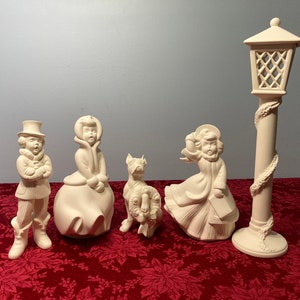 May include: A set of five white ceramic Christmas figurines, including a girl holding a book, a girl with a wreath, a boy singing, a girl in a coat, and a street lamp.