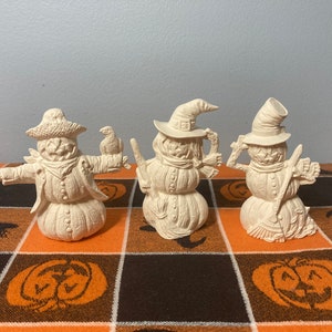 May include: Three unpainted ceramic scarecrow figurines. The scarecrows have pumpkin-shaped bodies and straw hats. One holds a bird, another a broom, and the third a mug. The figurines are on a Halloween-themed orange and black checkered tablecloth.
