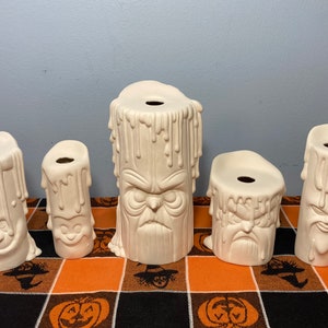 May include: Five white ceramic candle holders shaped like melting candles with spooky faces. The candle holders are arranged in a row on a checkered orange and black tablecloth.