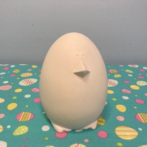 May include: White ceramic egg-shaped figurine with a beak and two eyes. The figurine is sitting on a turquoise background with a pattern of colorful eggs.