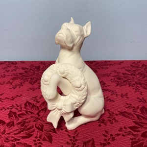 May include: White ceramic figurine of a boxer dog holding a Christmas wreath with a bow. The dog is sitting on a red background.