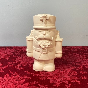 May include: A ceramic nutcracker figurine in a cream color. The figurine has a traditional military uniform design, complete with a hat, mustache, and decorative details. It stands on a red floral patterned surface.