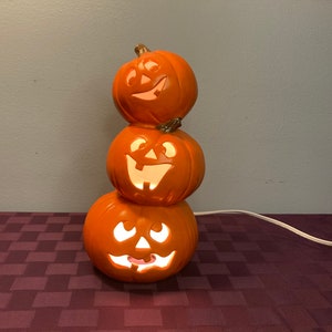 May include: A stack of three orange ceramic pumpkins with carved jack-o'-lantern faces. The pumpkins are lit from within, creating a warm glow.