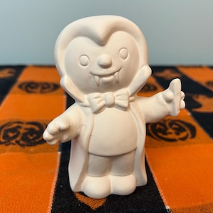 May include: A matte white ceramic figurine of a vampire character. The figure features a high-collared cape, a bow tie, and visible fangs. It is sculpted with rounded features, including a smooth head, small circular eyes, and a raised hand.