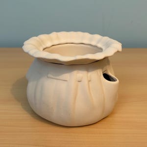 May include: A matte white ceramic vessel with a rounded, pleated body resembling a fabric pouch. The top edge features a wide, scalloped, ruffled rim. A small, circular opening is visible on the side of the body, integrated into the pleats.