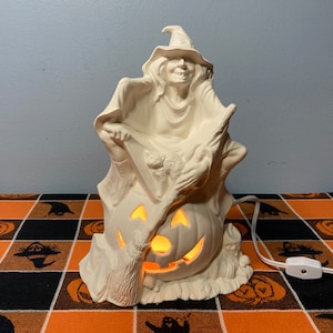 May include: White ceramic Halloween witch figurine holding a broom and sitting on a pumpkin. The witch is wearing a hat and has a long flowing robe. The pumpkin has a carved jack-o'-lantern face.