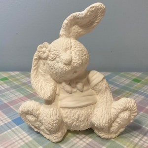 May include: White ceramic bunny figurine with a flower on its ear and a bow tie. The bunny is sitting with its ears up and its head tilted to the side.