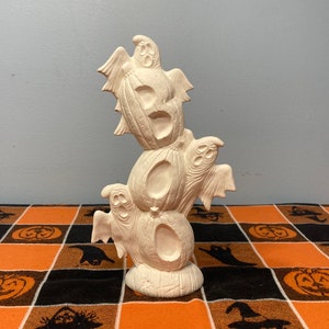 May include: A stacked Halloween decoration featuring three carved pumpkin shapes with ghost figures. The unpainted ceramic piece has a textured surface and is designed for seasonal display. The base is a rounded shape.
