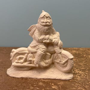 May include: A ceramic figurine of Santa Claus riding a motorcycle. The figure is painted in a neutral color, with details like a beard, goggles, and boots. The motorcycle has detailed wheels and a headlight.