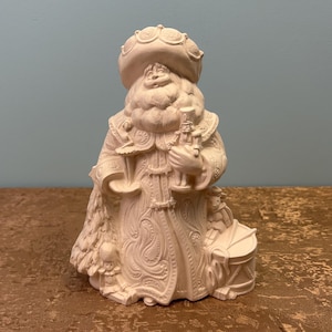 May include: A cream-colored figurine of a Santa Claus-like figure. The figure is wearing a hat and a long coat with intricate embossed designs. It holds a small bottle and a tray. A drum is at the base of the figure.