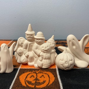 Puede incluir: Colecci&oacute;n de figuras de Halloween de cer&aacute;mica sin pintar. El conjunto incluye fantasmas, ni&ntilde;os que piden dulces y una calabaza. Las figuras est&aacute;n dispuestas sobre un mantel negro y naranja con tem&aacute;tica de Halloween.