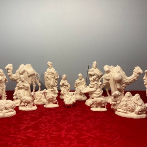 May include: A white ceramic nativity scene with figures of Mary, Joseph, baby Jesus, three wise men, shepherds, camels, and sheep. The figures are all standing on a red tablecloth.