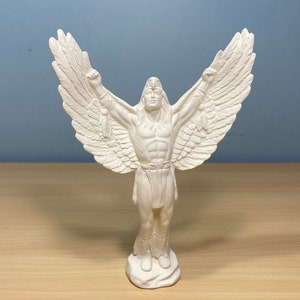May include: White ceramic statue of a person with outstretched arms and large wings. The person is wearing a feathered headdress and a skirt.