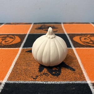 Puede incluir: Una peque&ntilde;a calabaza de cer&aacute;mica sin pintar con una superficie texturizada. La calabaza es de color blanquecino y se encuentra sobre un mantel a cuadros naranja, negro y blanco con tem&aacute;tica de Halloween con dise&ntilde;os de calabazas y murci&eacute;lagos.