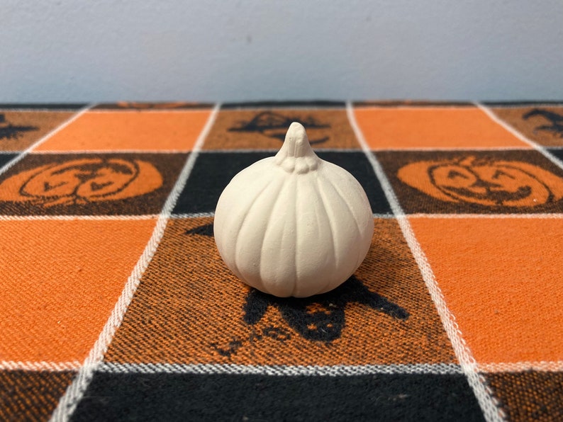 Puede incluir: Una peque&ntilde;a calabaza de cer&aacute;mica sin pintar con una superficie texturizada. La calabaza es de color blanquecino y se encuentra sobre un mantel a cuadros naranja, negro y blanco con tem&aacute;tica de Halloween con dise&ntilde;os de calabazas y murci&eacute;lagos.