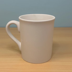 May include: A plain white ceramic mug with a curved handle. The mug is cylindrical with a slightly flared rim. It sits on a light-colored wooden surface against a light blue background. The mug is empty.