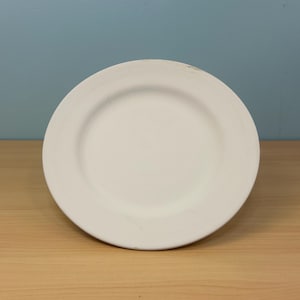 May include: A white ceramic plate with a wide rim. The plate is round and sits on a light brown wooden surface. The background is a solid light blue color.