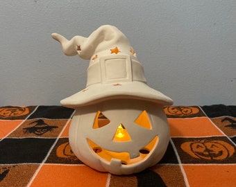 May include: White ceramic pumpkin with a witch's hat, featuring a carved jack-o'-lantern face and star cutouts. The pumpkin has a light inside, illuminating the face.