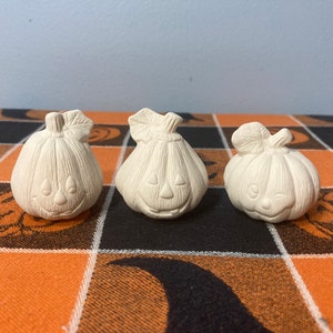 May include: Three white ceramic pumpkin figurines with painted facial features. The pumpkins are arranged in a row on a black and orange checkered tablecloth.