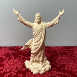 May include: White ceramic figurine of Jesus Christ with outstretched arms, standing on a cloud. The figurine is in a prayerful pose, with his eyes closed and his head tilted slightly upwards.