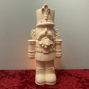 May include: A cream-coloured ceramic nutcracker figurine with a tall hat, curly moustache, and beard. The figurine has cylindrical arms and boots. Small holes allow light to shine through. The figurine is approximately 30 cm tall.