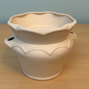 May include: A matte white ceramic wax warmer featuring a two-piece design with a scalloped rim on the top dish and a base with integrated side handles. The surface is smooth and unglazed, showcasing a clean, neutral aesthetic for home fragrance.