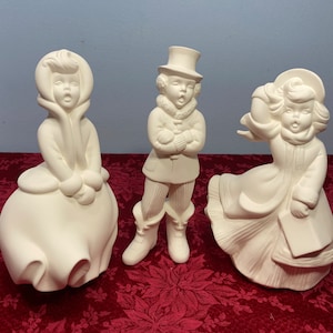 May include: Three white ceramic figurines of carolers. The figurines are in various poses, including a girl with her hands clasped in front of her, a boy with his hands clasped in front of him, and a girl holding a book. The figurines are all wearing traditional caroler clothing.