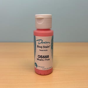 May include: A bottle of Duncan Bisq-Stain Opaque Acrylic paint in Miami Pink (OS558). The bottle has a white cap and a label with the brand name and product details. The bottle contains 2 US FL OZ (59 ml) of paint.