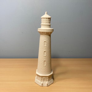 May include: A white ceramic lighthouse figurine with a detailed design. The lighthouse has a tall, cylindrical tower with three windows and a small, domed roof.