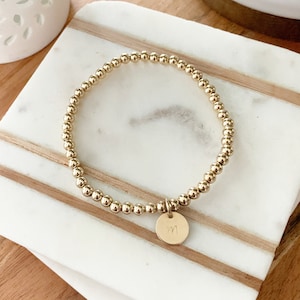 Könnte beinhalten: Ein goldfarbenes Perlenarmband mit einem kleinen runden Anhänger. Der Anhänger hat den Buchstaben "M" eingraviert.